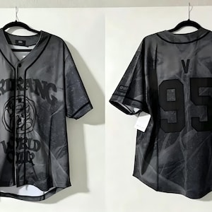 Peut inclure: Un maillot de baseball à manches courtes et boutonnage, dans des tons de gris et de noir. L'avant présente le texte ARIRANG WORLD TOUR avec un motif circulaire. Le dos affiche la lettre V au-dessus du nombre 95 en grands caractères noirs.