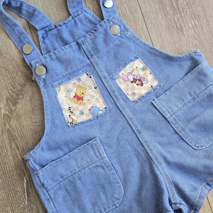 May include: Light blue denim baby overalls with Winnie the Pooh character patches on the front pockets. The overalls have adjustable straps and metal button closures. The patches feature Winnie the Pooh, Piglet, and Eeyore.
