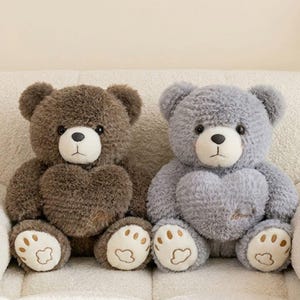 May include: Two plush teddy bears, one brown and one grey, sit side-by-side. Each bear has a heart-shaped chest and paw-shaped feet. The brown bear has light brown paw pads, while the grey bear has beige paw pads.
