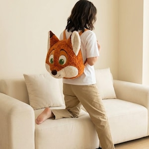 May include: A plush backpack shaped like a fox's head, featuring orange, white, and brown fur, with large green eyes and a black nose. The backpack has adjustable straps and is worn by a person. The fox backpack is a fun accessory.