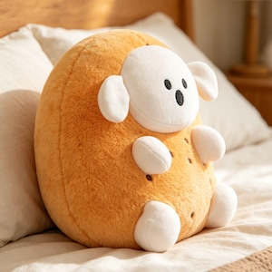 May include: A plush, round sheep toy with a light brown body and a white face, ears, and limbs. The sheep has a black nose and eyes. The toy is sitting on a bed with white pillows.