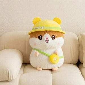 Pode incluir: Um brinquedo de pelúcia de hamster com um chapéu amarelo e uma alça verde. O brinquedo tem pelo marrom e branco, olhos grandes e pretos e um nariz rosa. Está sentado em um sofá creme.