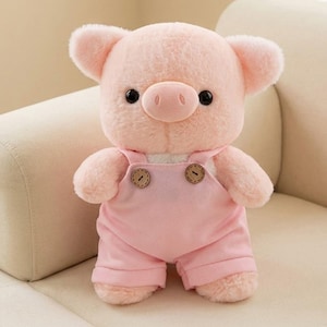 Puede incluir: Un peluche rosa con forma de cerdo, con grandes ojos negros, un hocico redondo y orejas pequeñas. El cerdo lleva un peto rosa con dos botones marrones. El juguete es suave y tierno, perfecto para niños.