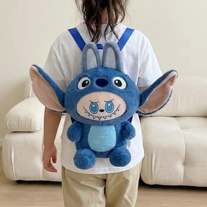 Könnte beinhalten: Ein blauer Plüschrucksack in Form einer Zeichentrickfigur mit großen Ohren und einem lächelnden Gesicht. Der Rucksack hat verstellbare Träger und wird von einer Person getragen, die ein weißes T-Shirt und eine khakifarbene Hose trägt.