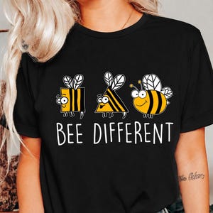 Puede incluir: Una camiseta negra de manga corta con un estampado gráfico de tres abejas de dibujos animados en formas cuadrada, triangular y redonda. Debajo de las abejas, el texto blanco BEE DIFFERENT aparece en una fuente clara y sin remates.