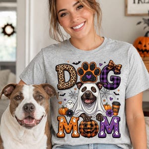 Könnte beinhalten: Ein hellgraues T-Shirt mit einem Grafikdruck und dem Text DOG MOM. Das Design zeigt einen Hund im Geisterkostüm, einen Pfotenabdruck, einen Kürbis, einen Kaffeebecher, Fledermäuse, Spinnen und Spinnweben in Orange und Lila.