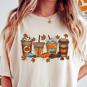Könnte beinhalten: Ein cremefarbenes T-Shirt mit einem Grafikdruck von vier Kaffeetassen, die mit herbstlichen Elementen wie orangefarbenen und blauen Kürbissen, Tannenzapfen, Eicheln und verstreuten Ahornblättern in Gelb-, Orange- und Brauntönen verziert sind.