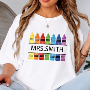 Könnte beinhalten: Ein weißes T-Shirt mit einer Grafik von sieben bunten Buntstiften in einer Reihe. Der Text MRS.SMITH ist in schwarzen Großbuchstaben über die Mitte der Stifte gedruckt. Die Stifte sind gelb, orange, rot, grün, blau, indigo und violett.