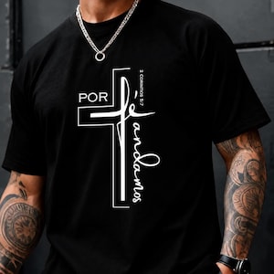 Peut inclure: Un t-shirt noir à manches courtes avec un motif graphique blanc représentant une croix. Le texte "POR" est imprimé au-dessus de la croix, tandis que "fe andamos" est écrit en cursive le long de la barre verticale. "2 CORINTIOS 5:7" est présent.