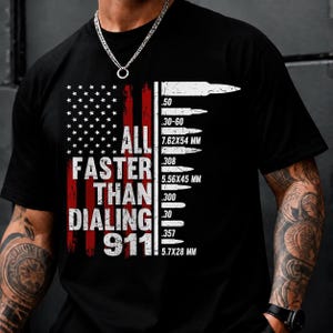 Pode incluir: Uma t-shirt preta de manga curta com um gráfico de uma bandeira americana desgastada em vermelho e branco. O texto diz ALL FASTER THAN DIALING 911, acompanhado por uma lista vertical de ilustrações de balas com calibres como .50, .30-60 e .308.