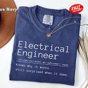 May include: A true navy blue t-shirt with the words "Electrical Engineer" printed in white. The definition of the term is included below the title. A gold necklace with a pendant is draped across the shirt. A "Fast Delivery" sticker is visible.