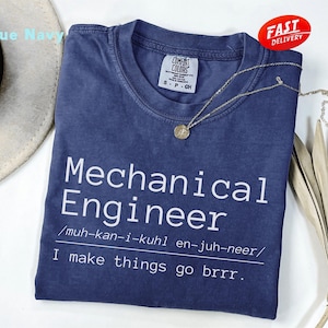 May include: A navy blue short-sleeved t-shirt with white text that reads Mechanical Engineer, followed by a phonetic pronunciation guide and the phrase I make things go brrr. The shirt features a crew neckline and a Comfort Colors brand label.