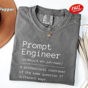 May include: A gray t-shirt with the text "Prompt Engineer" in white. The shirt includes a definition and pronunciation guide. A gold necklace and a "Fast Delivery" sticker are also visible.