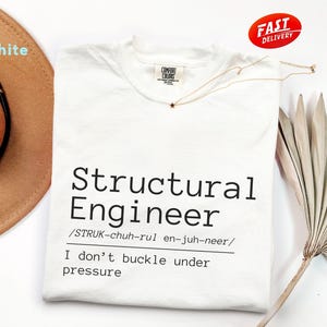 May include: A white short-sleeved t-shirt featuring the black printed text Structural Engineer, its phonetic pronunciation, and the phrase I don't buckle under pressure. The shirt has a Comfort Colors brand label at the inner neckline.
