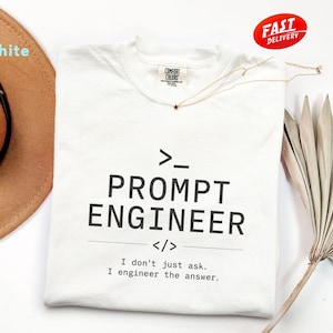 May include: White t-shirt with the text "PROMPT ENGINEER" in black, along with the phrase "I don't just ask. I engineer the answer." A gold necklace with a star pendant is on the shirt. A brown hat and a dried plant are also in the image.