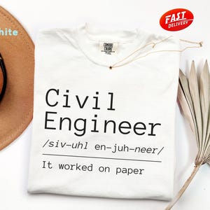 May include: White t-shirt with the text "Civil Engineer" in black, including a phonetic pronunciation. The phrase "It worked on paper" is below the title. A gold necklace with a star pendant is on the shirt.