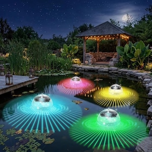 Puede incluir: Escena nocturna de un estanque iluminado por cuatro luces solares flotantes. Cada luz emite un color diferente: azul, rosa, amarillo y verde. Las luces están rodeadas de rayos. Un cenador de madera y la luna llena se ven al fondo.