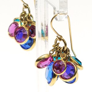 May include: A pair of dangle earrings featuring clusters of faceted round crystals in vibrant shades of magenta, purple, and bright blue. The crystals are set in gold-toned metal settings and hang from simple gold-toned curved ear wires.