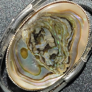 Könnte beinhalten: Vintage ovale Brosche mit einem Zentrum aus Abalone-Muschel. Die Muschel zeigt schillernde Farben von Grün, Blau und Gold, eingefasst in einen silberfarbenen Metallrahmen. Der Rahmen hat ein mehrschichtiges Design.
