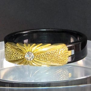 May include: A black band ring featuring a central gold-toned rectangular accent with a sunburst pattern, textured stippled sides, and a single clear round rhinestone set in the middle of the gold-toned metal surface.