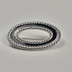 May include: An oval-shaped brooch featuring a polished silver-tone twisted rope border surrounding a smooth, glossy black enamel inlay. The center of the brooch is open, revealing a thin silver pin mechanism attached to the back of the frame.