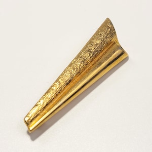 May include: A gold-toned metal brooch shaped like an elongated triangle. The surface features a distinct contrast between a rough, textured side and a smooth, polished side, meeting at a central ridge that runs the length of the piece.