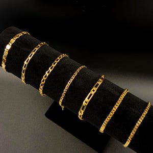Puede incluir: Una colección de seis pulseras de cadena de metal en tono dorado exhibidas en un soporte cilíndrico negro. Las pulseras presentan varios estilos de eslabones, incluyendo eslabones tipo cable, barbados y ovalados alargados.