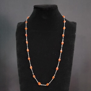May include: A long necklace featuring a series of small, smooth, oval-shaped orange carnelian beads spaced out by thin, polished silver-toned metal bars. The beads are arranged in a repeating pattern along the delicate silver-toned metal chain.