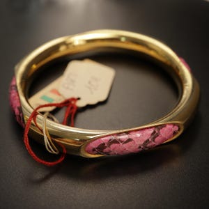 May include: A gold-toned bangle bracelet featuring inlaid sections of pink and black snakeskin-patterned material. A small, light-colored rectangular paper tag with handwritten text is attached to the bracelet by a thin red string.