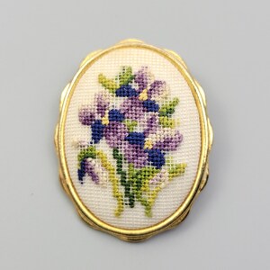 May include: An oval brooch featuring a gold-toned metal frame with a scalloped edge. The center contains a piece of white fabric with a cross-stitch embroidery design of a bouquet of purple, blue, and green flowers on a grid pattern.