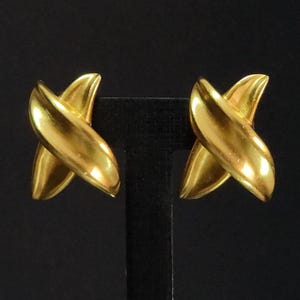 May include: A pair of gold-toned earrings featuring a polished, smooth X-shaped design. Each earring consists of two curved, intersecting bands that create a symmetrical, dimensional form with a reflective metallic finish and rounded edges.