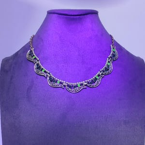 May include: A silver-toned necklace with a scalloped design, featuring dark blue and green gemstones. The necklace is displayed on a purple mannequin, showcasing its intricate details and elegant style. The necklace has a delicate chain.