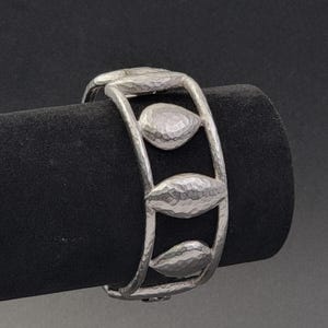 Può includere: Un bracciale rigido color argento con una texture martellata. Il design è composto da due fasce parallele collegate da tre elementi distinti a forma di foglia, ognuno dei quali presenta una finitura martellata sulla superficie.