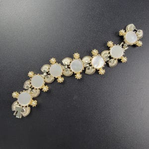Pode incluir: Uma pulseira de elos em tom dourado com seis cabochões circulares de madrepérola branca. Cada elo é acentuado com pequenas pérolas falsas redondas de cor creme e detalhes metálicos texturizados em forma de leque com fecho.