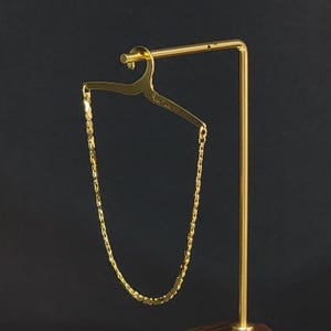May include: A gold-toned metal chain necklace suspended from a miniature gold-toned metal clothes hanger display stand. The hanger features a small hook at the top and a flat horizontal bar with engraved text that reads Pierre Cardin Paris.