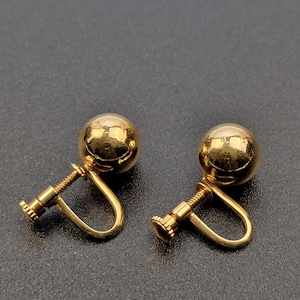 May include: A pair of gold-toned screw-back earrings featuring smooth, spherical ball studs. Each earring has a polished, reflective surface and a threaded screw mechanism designed for non-pierced ears, showcasing a classic, rounded design.