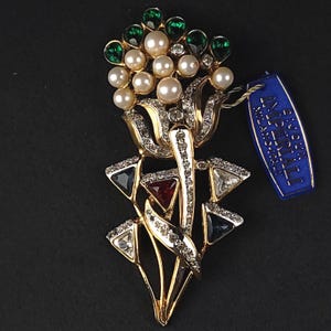 May include: A gold-tone brooch shaped like a flower bouquet, featuring white faux pearls, emerald green teardrop stones, and clear, red, and dark blue triangular crystals. A blue tag attached reads BIJOUX IMPERIALI MILANO ITALY.