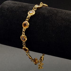 Op de afbeelding: Een goudkleurige kettingarmband met een reeks ronde, barnsteenkleurige gefacetteerde stenen in metalen zettingen. De armband is voorzien van een kleine, bungelende metalen bedel in de vorm van een Scottie-hond bij de karabijnsluiting.
