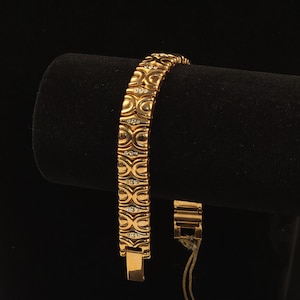 Puede incluir: Una pulsera de eslabones en tono dorado con un patrón repetitivo de formas curvas similares a lazos. Cada eslabón está acentuado con una pequeña piedra redonda y transparente incrustada en el centro. La pulsera tiene un acabado metálico y cierre.