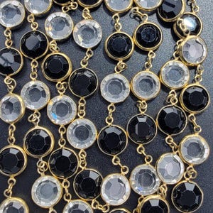 Puede incluir: Un collar de múltiples hilos con piedras circulares facetadas, alternando entre transparentes y negras, engastadas en metal dorado. Las piedras están unidas en filas verticales, creando un patrón repetitivo de elementos circulares claros y oscuros.