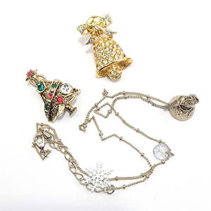 May include: A gold-tone bell brooch encrusted with clear rhinestones and topped with a bow, a gold-tone Christmas tree brooch with red and green rhinestones, and a gold-tone chain necklace featuring a white snowflake charm and a small bell pendant.