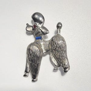 Op de afbeelding: Een zilverkleurige poedelbroche met een getextureerde metalen afwerking die vacht nabootst. De hondenfiguur is versierd met een kleine, rechthoekige blauwe edelsteen als halsband en een minuscuul, rond rood edelsteenaccent bij de nek.