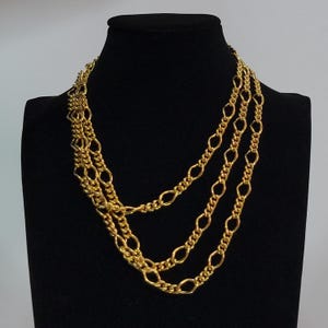 Op de afbeelding: Een goudkleurige ketting met meerdere strengen, bestaande uit een combinatie van langwerpige ovale schakels en getextureerde, strak geweven kettingsegmenten. Het ontwerp bestaat uit verschillende gelaagde lussen van variërende lengtes.