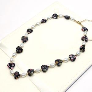 May include: A necklace featuring alternating black heart-shaped beads with colorful floral patterns and white oval pearls. The beads are separated by small gold-toned metal spacers, and the piece includes a gold-toned metal clasp and an adjustable chain.