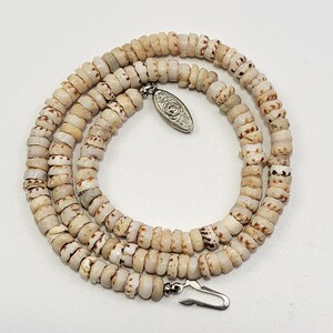 Puede incluir: Un collar compuesto por numerosas cuentas de concha pequeñas, circulares y de color blanco roto con patrones de motas marrones. La hebra presenta un dije ovalado de metal plateado con un diseño en relieve y un cierre de gancho de metal.