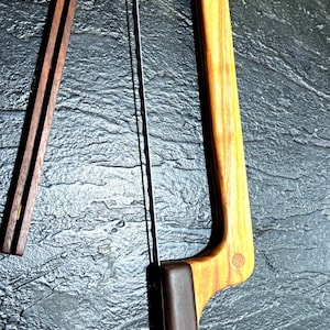 May include: A wooden bow saw with a dark brown leather handle and a thin metal blade. The saw has a light brown wooden frame with a decorative circular inlay. A separate wooden piece is also present.