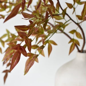 May include: A vase with a textured white finish holds a sprig of artificial foliage with red-brown leaves.