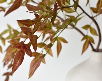 May include: A vase with a textured white finish holds a sprig of artificial foliage with red-brown leaves.