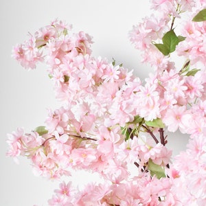 Puede incluir: Primer plano de una rama de flores de cerezo artificiales. Las flores son de color rosa suave, con m&uacute;ltiples capas de p&eacute;talos. Las ramas son marrones y hay algunas hojas verdes.