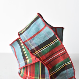 May include: A decorative ribbon featuring a plaid pattern in shades of light blue, forest green, black, and red. The fabric has a distinct woven texture with thin yellow and white accent stripes running throughout. The edges are finished with red stitching.